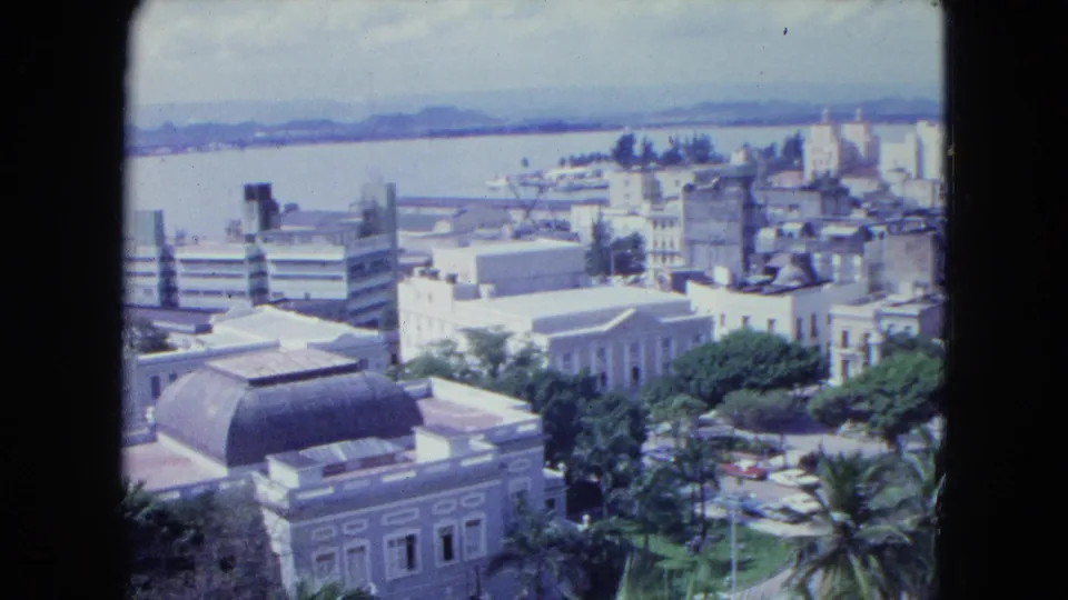 1964 San Juan panorama from above, showing waterfront buildings, tree-lined streets, parked cars, palms, water, and hills.