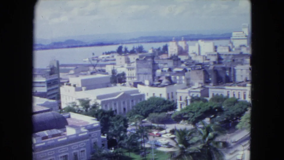 San Juan in 1964 seen from a rooftop or high overlook, with mid-century buildings, leafy streets, water, and distant hills.