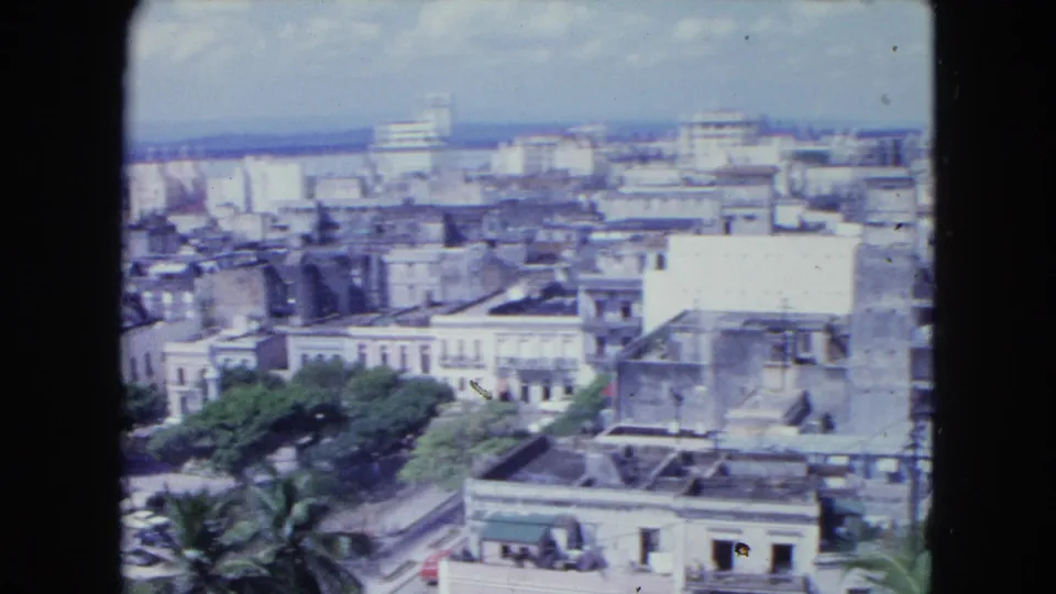1964 8mm frame from San Juan showing flat-roofed buildings, older facades, palms, and distant modern blocks.