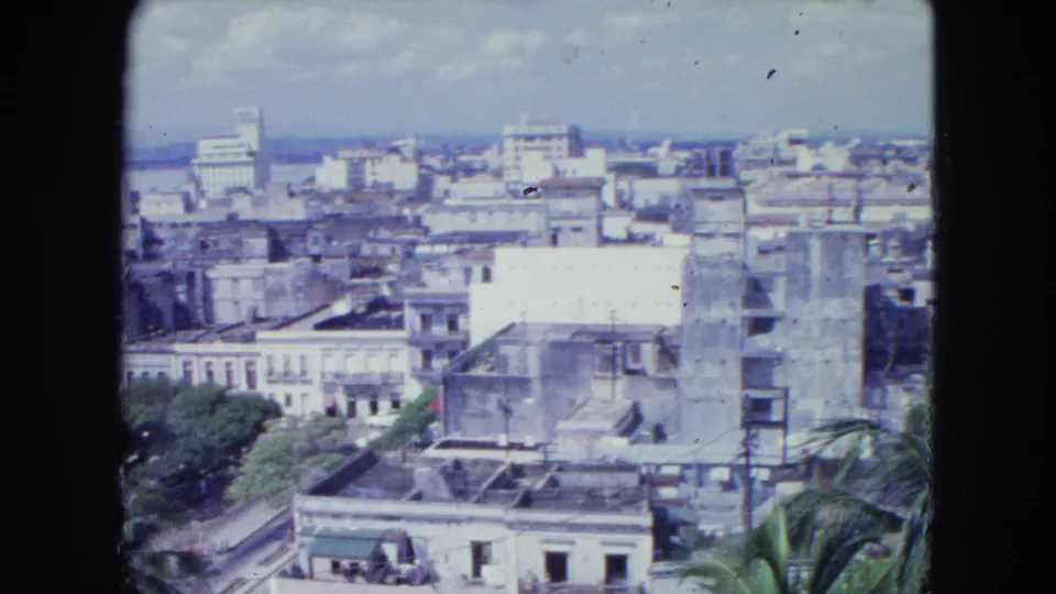 1964 San Juan panorama frame with layered rooftops, palms at the edge, a bright wall, and distant waterfront skyline.