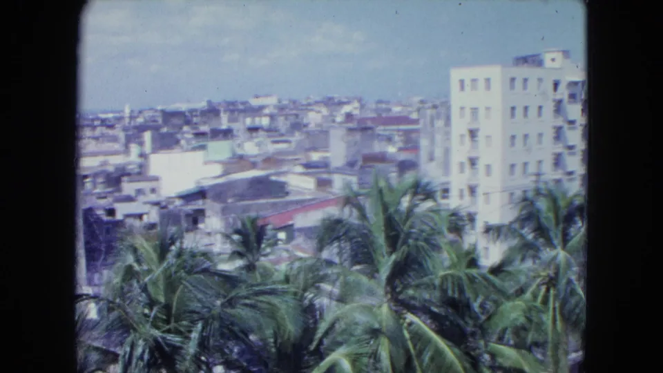1964 San Juan home-movie view with palms, dense rooftops, a white high-rise, and the ocean horizon beyond.
