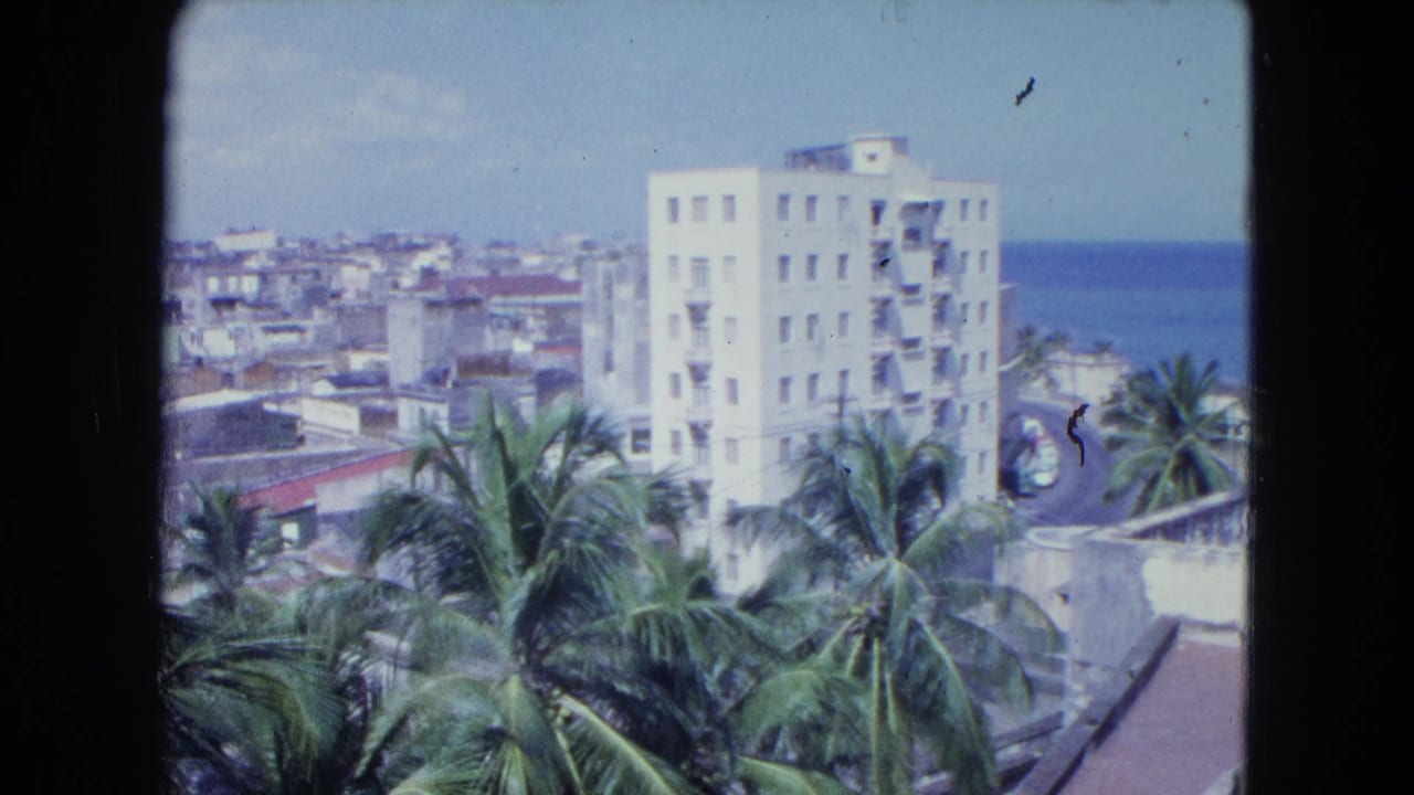 1964 San Juan 8mm frame showing palms below a white high-rise, low rooftops, and blue ocean to the right.