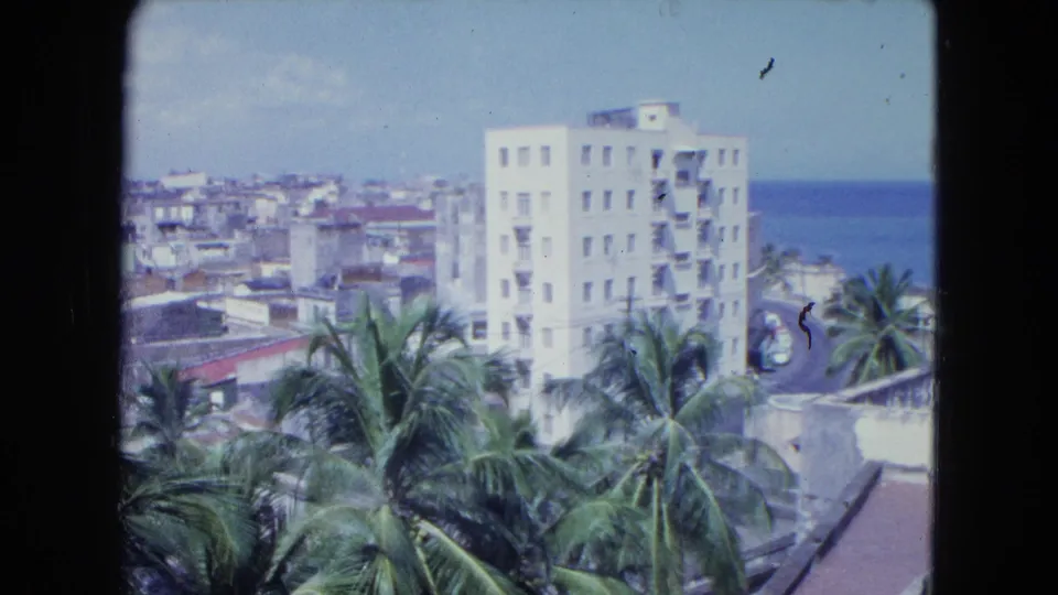 1964 San Juan 8mm frame showing palms below a white high-rise, low rooftops, and blue ocean to the right.