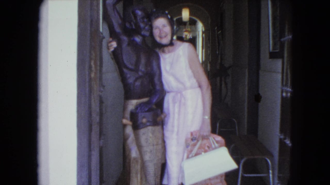 In 1964 San Juan home-movie footage, a woman in a pink dress smiles beside a dark bronze statue while holding a handbag and white item.