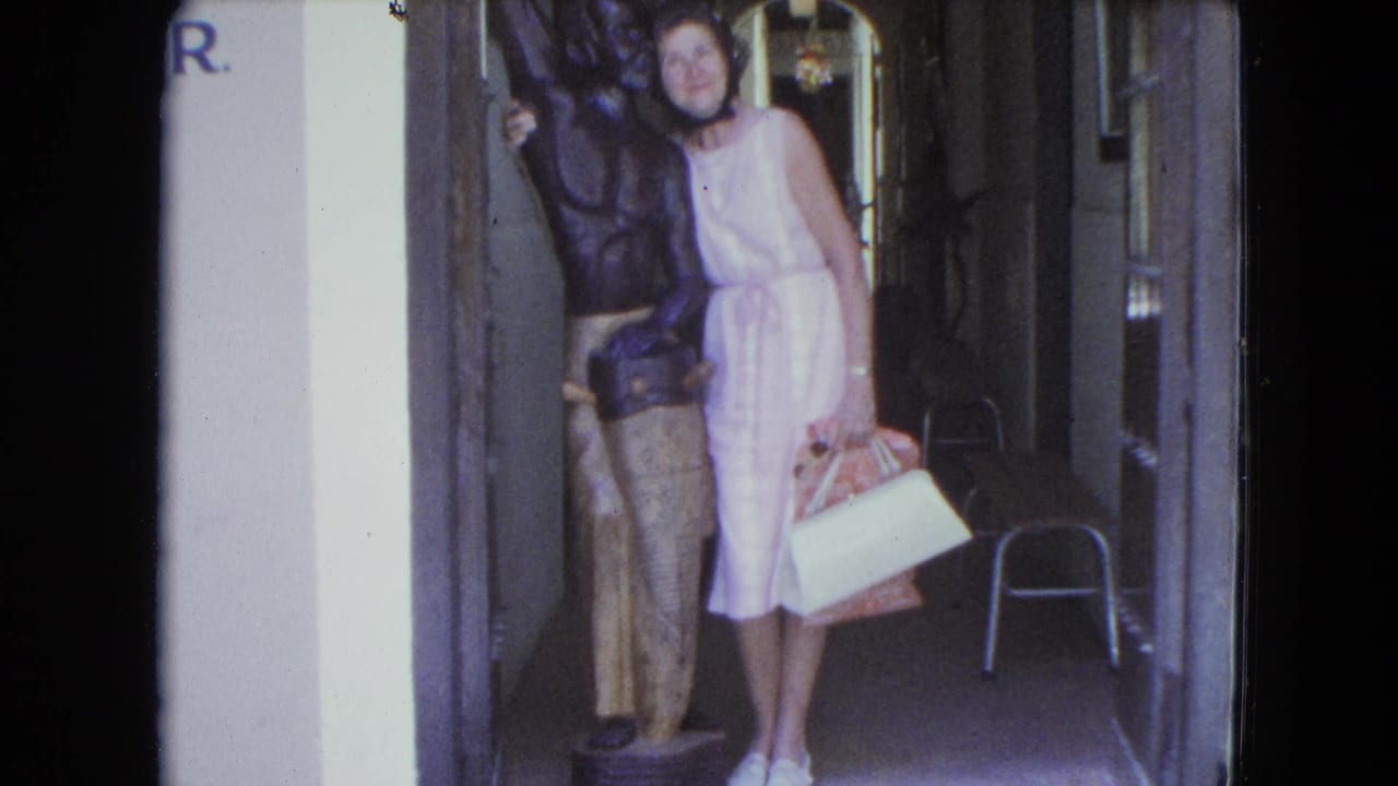 1964 San Juan home-movie frame showing a woman in a light pink dress posing next to a dark carved statue indoors.