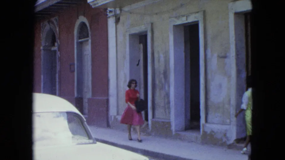 1964 San Juan frame of a woman carrying a dark bag while walking past stucco buildings and a white car.