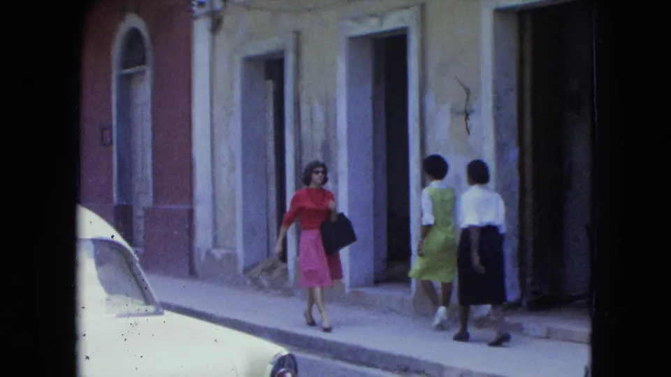1964 San Juan street frame with a woman in red and pink near two passing women and colonial-style building fronts.