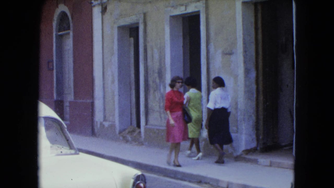 Three women walk on a sidewalk beside a white parked car and weathered buildings.
