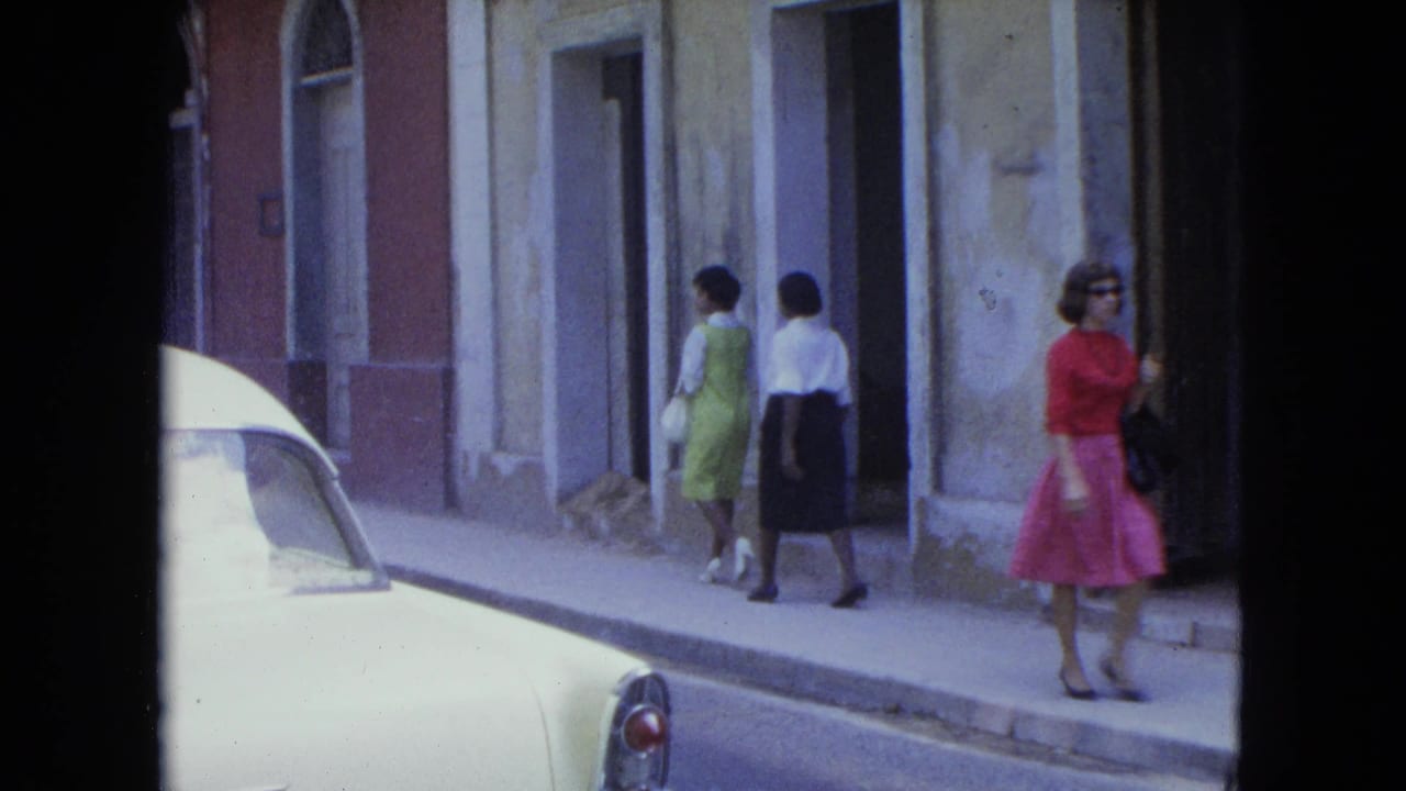 Women continue along the sidewalk as a woman in pink stands near a doorway.