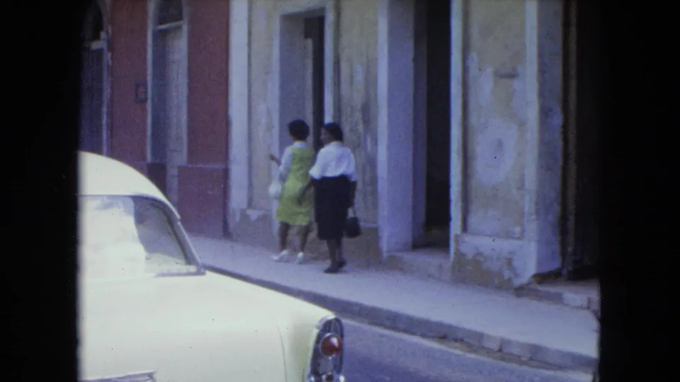 1964 San Juan street frame showing two women walking away along a sidewalk beside a parked white car.