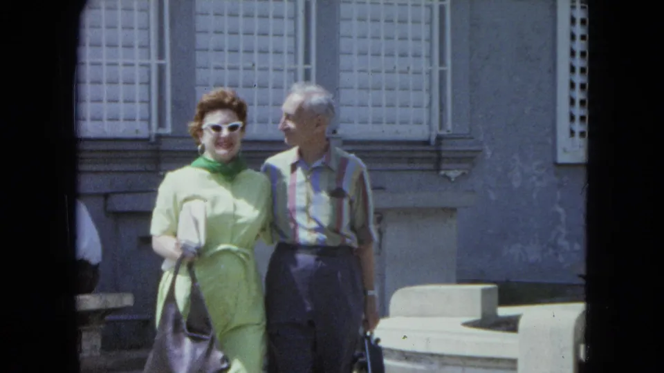A couple walks together in San Juan, with barred windows and a parked white car behind them.