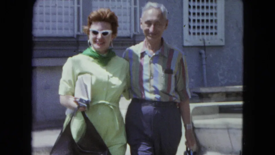 The couple remains close to the camera, the woman holding a dark handbag against a sunlit San Juan building exterior.