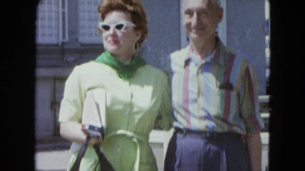 In 1964 San Juan home-movie footage, a woman in green with white sunglasses stands beside a man in a striped shirt outdoors.