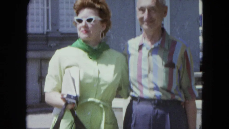 In 1964 San Juan home-movie footage, a woman in green with white sunglasses stands beside a man in a striped shirt outdoors.