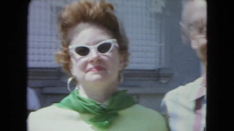 A close home-movie frame shows the woman in green with white cat-eye sunglasses and hoop earrings, with the man partly at right.