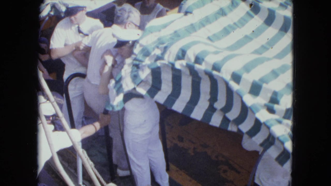 A man stands on a small boat while sailors gather under a striped canopy.