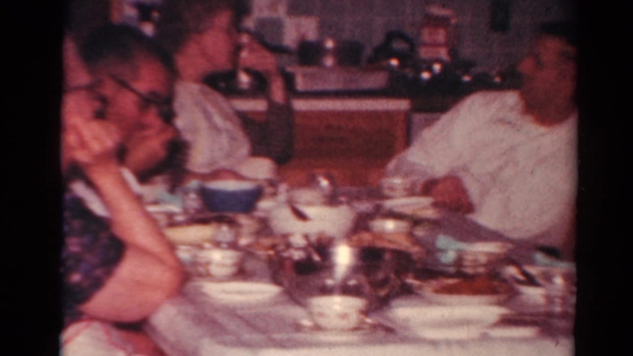 1964 Toledo home-movie frame showing adults gathered at a dining table with bowls, cups, and serving dishes in a kitchen interior.