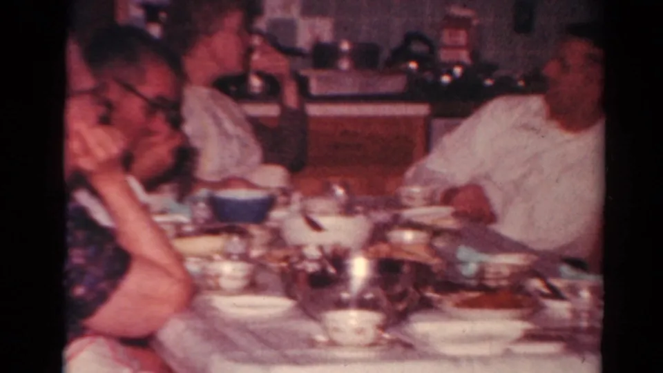 1964 Toledo home-movie frame showing adults gathered at a dining table with bowls, cups, and serving dishes in a kitchen interior.