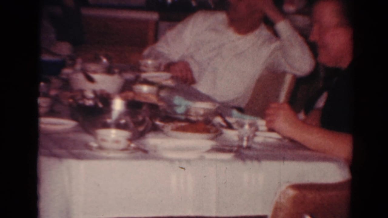 A man in a white shirt sits at a cluttered dining table during a 1964 Toledo family meal, with another adult partly visible nearby.