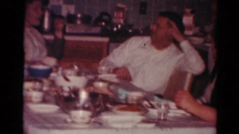 Adults remain seated around a crowded dining table in a private Toledo home interior, captured with soft 8mm color and grain.
