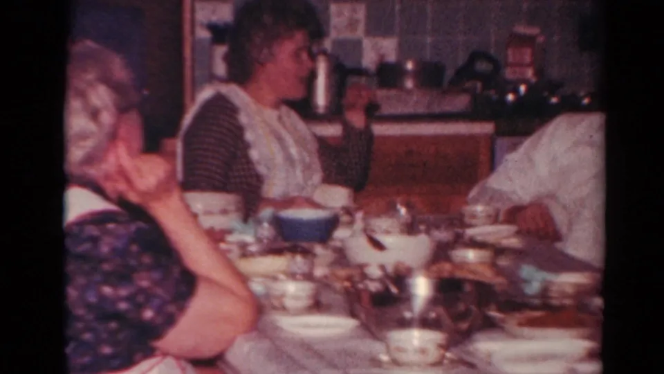 A 1964 Toledo home-movie frame shows several adults at a dining table, with kitchen counters and household objects behind them.