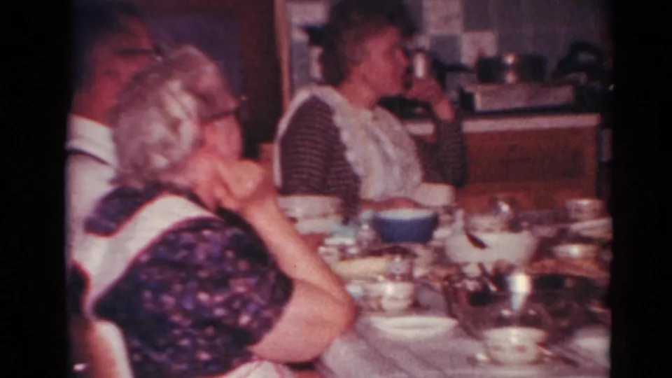 1964 Toledo home-movie frame of adults gathered around a kitchen table with bowls, cups, and serving dishes.