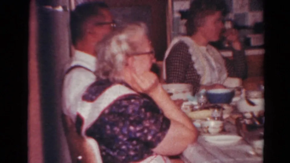 1964 Toledo home-movie frame showing adults in conversation around a kitchen table set with tableware.
