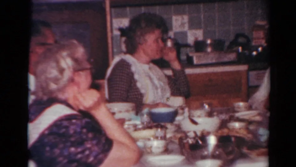 1964 Toledo home-movie frame of a family meal in a private kitchen, with adults seated beside bowls and cups.