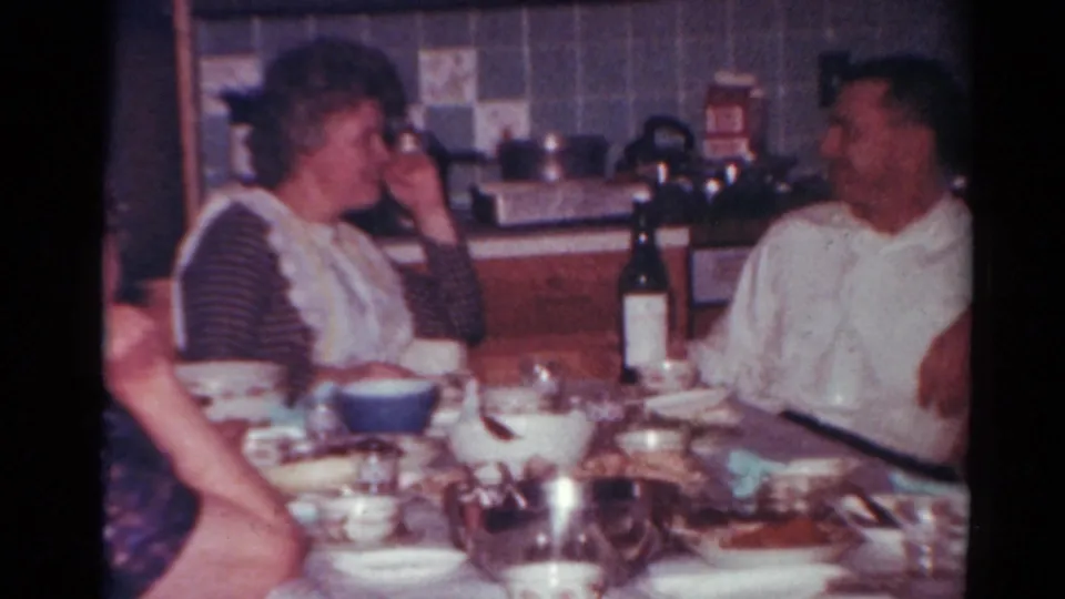 1964 Toledo home-movie frame showing adults conversing at a kitchen table with a bottle, bowls, and plates visible.