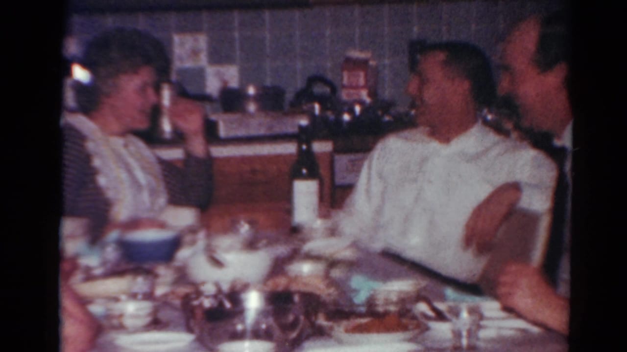 In a Toledo, Ohio 1964 home-movie frame, adults sit around a crowded dinner table while a woman gestures near a dark wine bottle.
