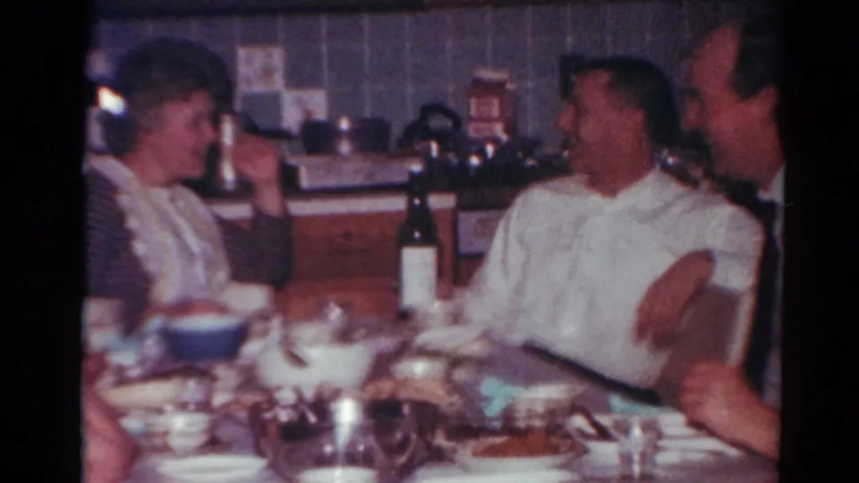 In a Toledo, Ohio 1964 home-movie frame, adults sit around a crowded dinner table while a woman gestures near a dark wine bottle.
