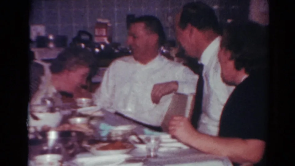 In the 1964 Toledo kitchen scene, adults lean and laugh around a holiday table with dishes, glasses, and a wine bottle visible.