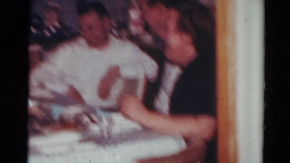 A soft, blurred 1964 Toledo home-movie frame shows adults at a dinner table, including a man in a dark shirt close to the camera.