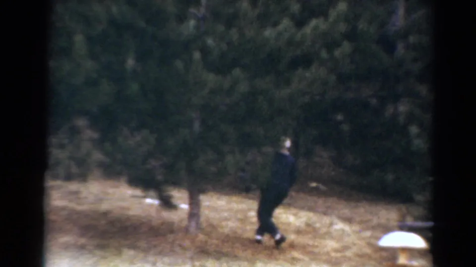 1964 Toledo frame with a dark-clothed figure moving through a wooded yard, partly softened by 8mm focus and grain.