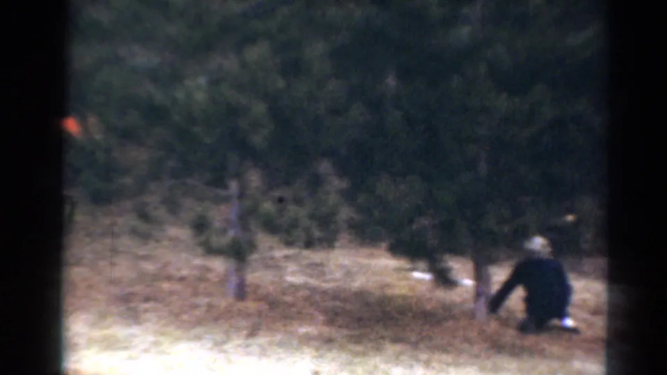 1964 Toledo home-movie frame showing a figure crouched low near trees during outdoor play in a wooded yard.