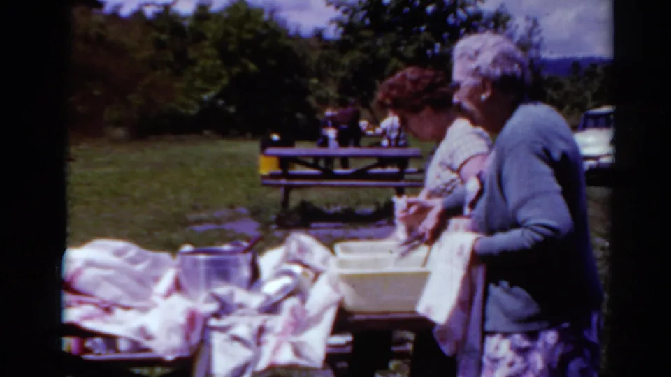 A middle-aged woman and older woman lean over a picnic table with cookware, towels, and a yellow dish basin in 1964 Toledo footage.