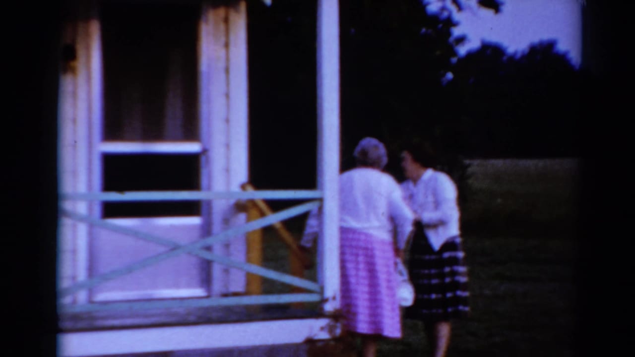 In 1964 Toledo home-movie footage, an older woman in pink and a younger woman in a plaid skirt stand together by a small house porch.