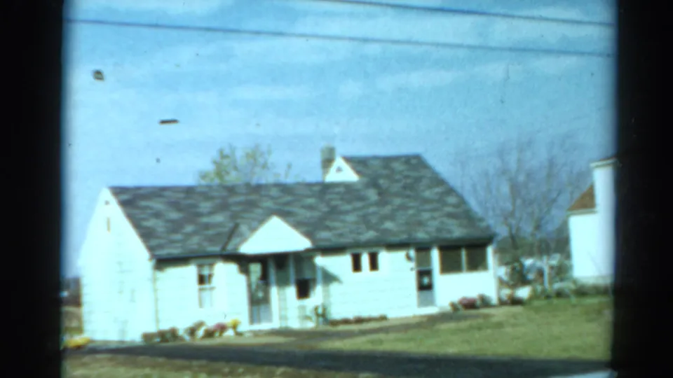 A small white suburban house appears across the lawn, with porch details, roofline, shrubs, and neighboring structures visible.