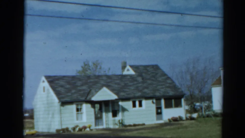 The clip ends on another distant view of the modest white house, lawn, porch area, bare trees, and adjacent building edge.