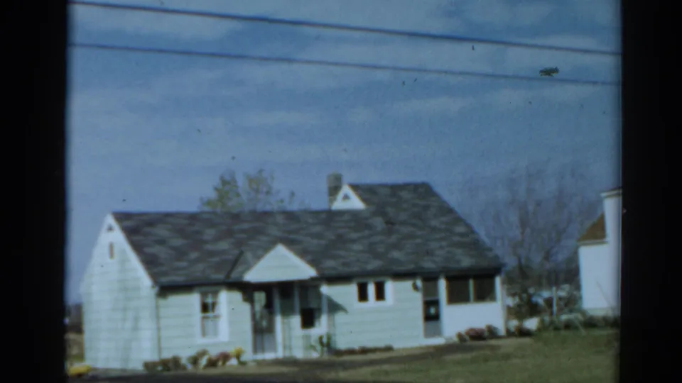 1964 Toledo home-movie frame showing a pale suburban house with a dark shingle roof, front porch, white trim and spring flowers.
