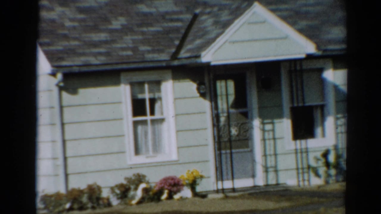 1964 Toledo home-movie frame showing a modest light-sided house with porch, window, roofline, walkway, and front flowers.