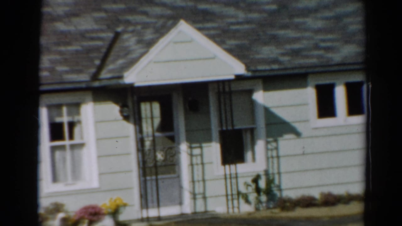 House front with porch, windows, flower bed, walkway, and dark clothes hanging near the entrance.