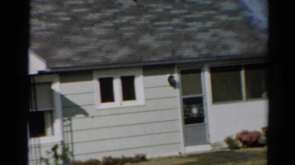 1964 Toledo home-movie frame showing the side portion of a modest house, including roofline, small windows, door, and garden flowers.