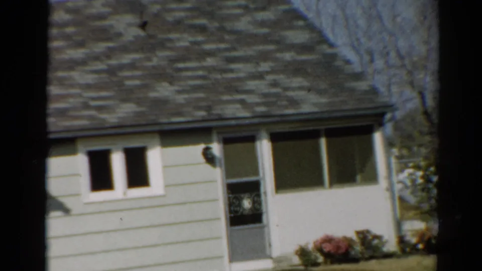 1964 Toledo frame showing a light-sided house exterior with dark roof, small windows, front door, flower bed, and bare branches.