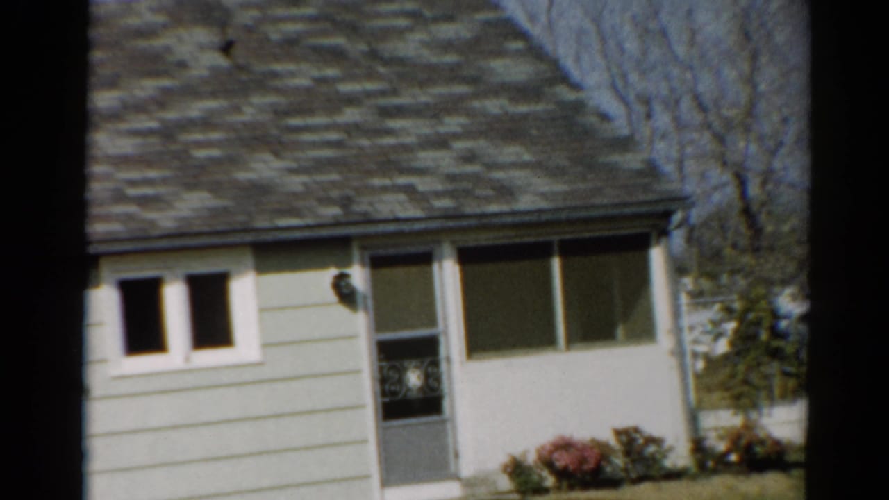 Front of a light-colored one-story house with white trim, screen door, picture window, lawn, and shrubs.