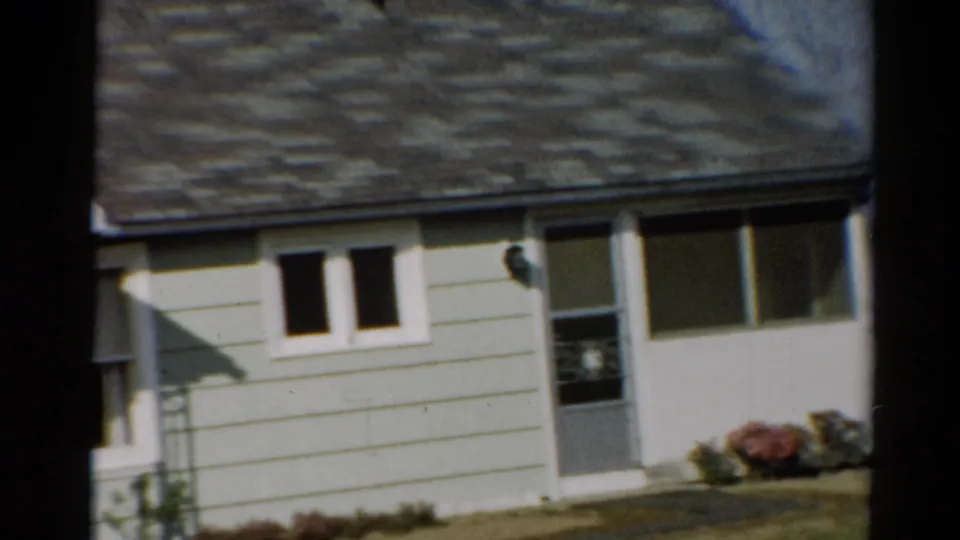 1964 Toledo frame showing the bungalow front during the pan, with porch edge at left, white-trimmed windows, screen door, lawn, and flower beds.