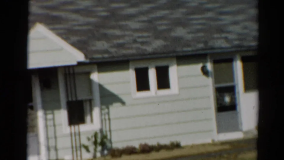 1964 Toledo frame with the porch side and railing visible beside the light-colored bungalow, plus small windows, screen door, lawn, and low plants.