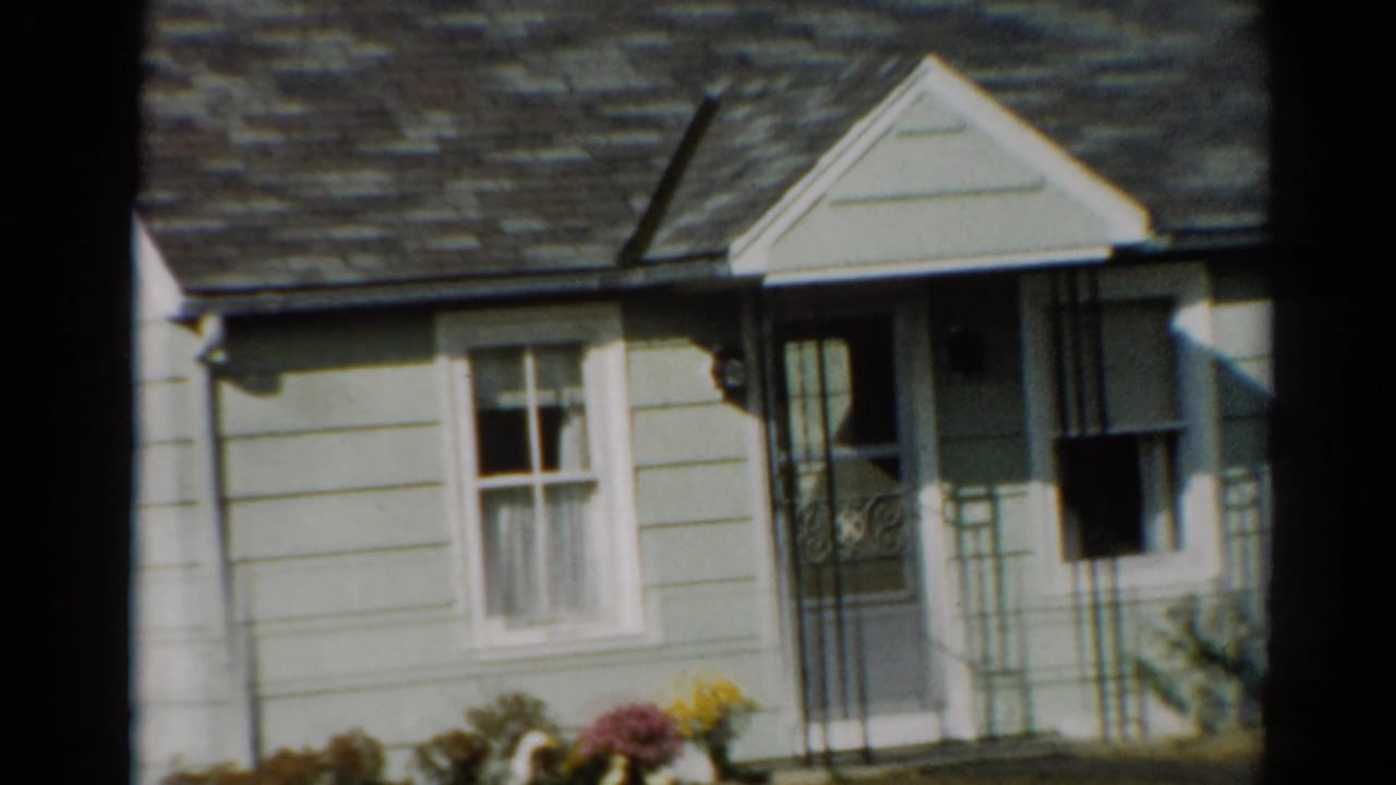 Pale green clapboard house with white trim, front door, window, porch rails, and flower bed.