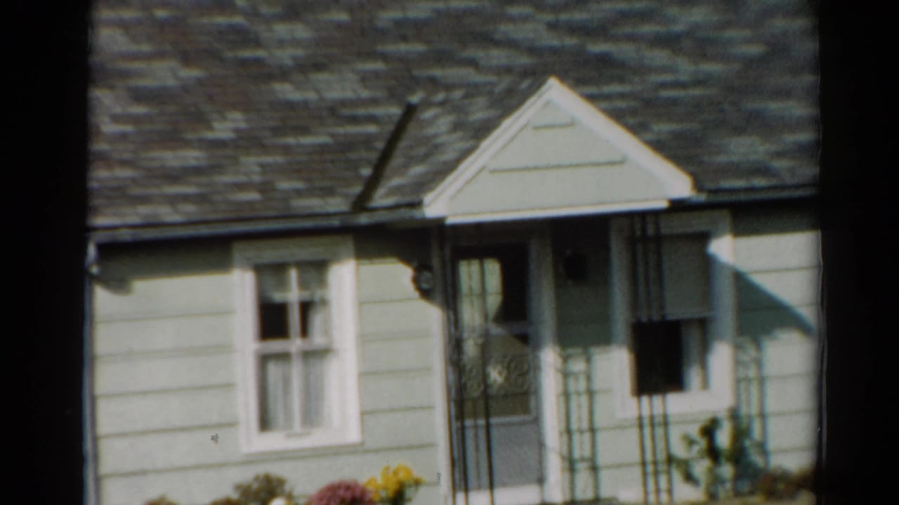 1964 Toledo frame of a sunlit suburban house front with clapboard siding, curtained window, porch entrance, and decorative railing.