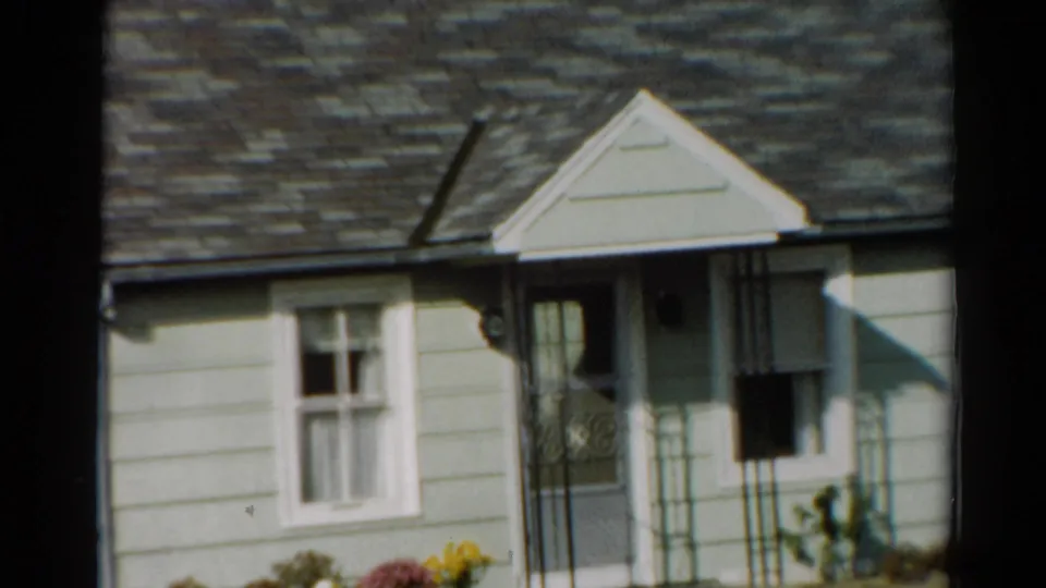 1964 Toledo frame of a sunlit suburban house front with clapboard siding, curtained window, porch entrance, and decorative railing.